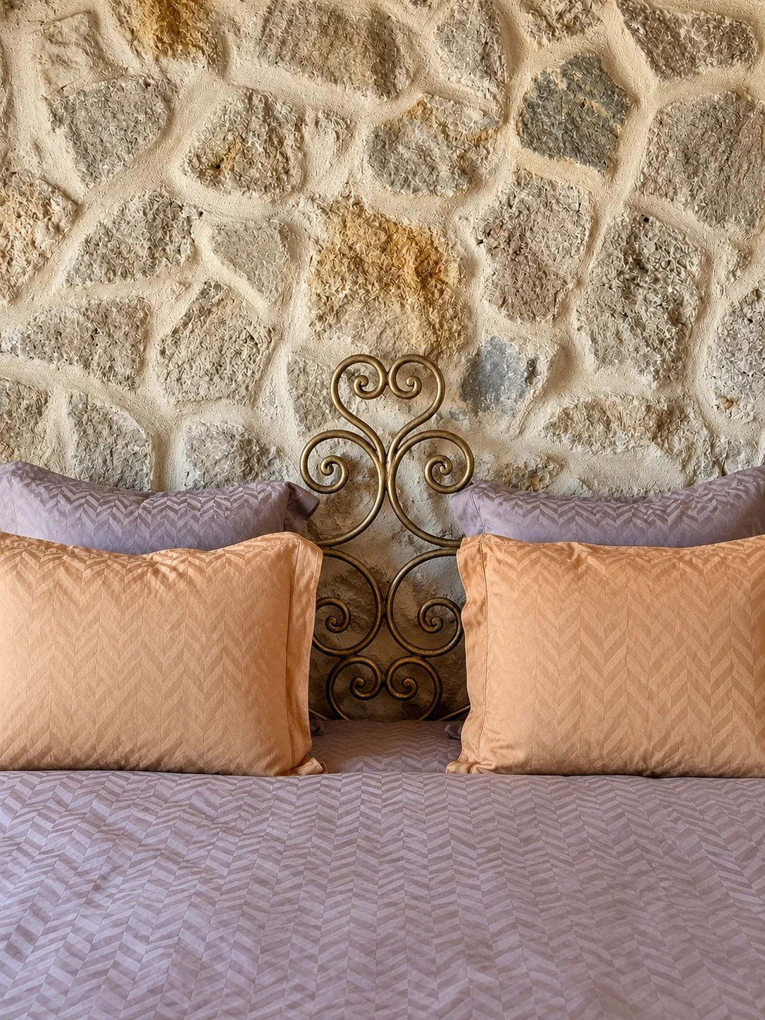 Lit de la Suite Deluxe avec tête de lit en fer forgé et linge mauve, devant un mur de pierre à L'Escondida